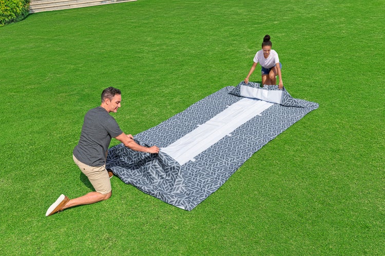 Ein Mann und eine Frau entfalten eine gemusterte Pool-Solarfolie auf einer Rasenfläche im Garten.