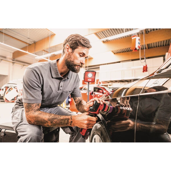 Ein Handwerker poliert mit einem Akku-Exzenterschleifer die Karosserie eines schwarzen Autos in einer hellen Werkstatt.