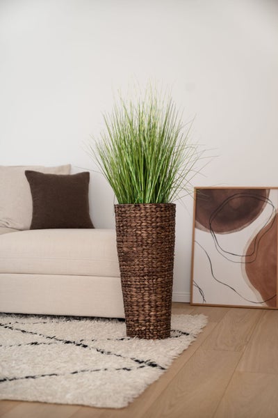 Künstliches Ziergras in einem hohen braunen geflochtenen Übertopf in einem hellen Wohnzimmer neben einem Sofa.