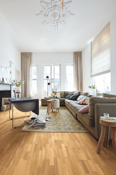Helles Wohnzimmer mit Eichenparkett, großem Ecksofa, grauem Sessel, gemustertem Teppich und hohen Fenstern.