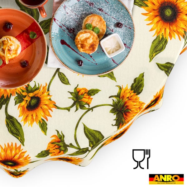 Abwaschbare Tischdecke mit Sonnenblumen-Muster auf einem gedeckten Tisch. Mit Lebensmittelecht-Symbol und ANRO Logo.