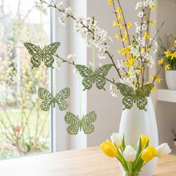 Osterdekoration zum Aufhängen in Form von ausgestanzten Schmetterlingen an blühenden Zweigen in einer Vase mit Tulpen auf einem Holztisch.