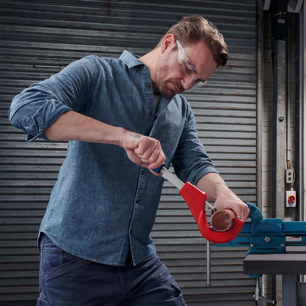Ein Mann mit Schutzbrille nutzt einen roten Rohrabschneider zum Trennen eines in einem Schraubstock fixierten Kupferrohrs.