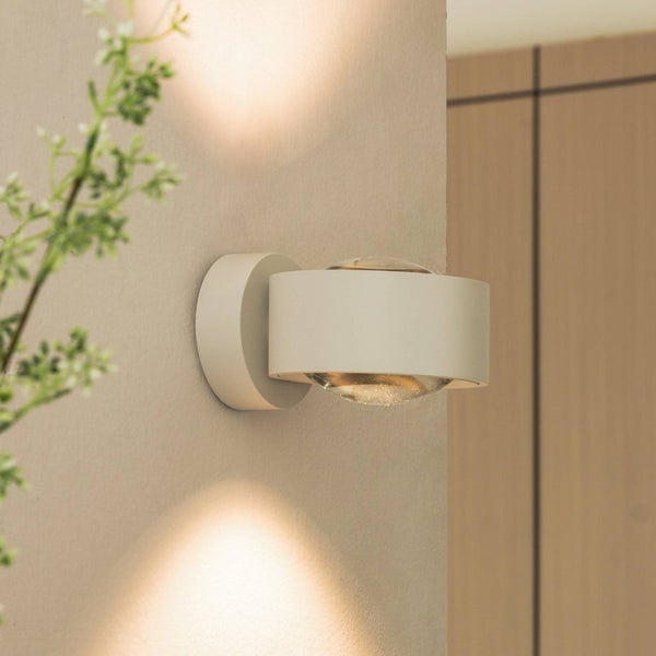 Moderne Wandleuchte mit Lichtkegeln nach oben und unten an einer strukturierten Wand, rundes Metallgehäuse, warmweißes Licht, helle Atmosphäre.