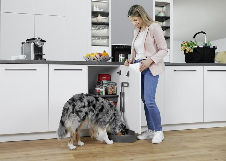 Moderne weiße Küche mit einer Frau und einem Hund, der aus einem an der Schranktür montierten Futternapf frisst.