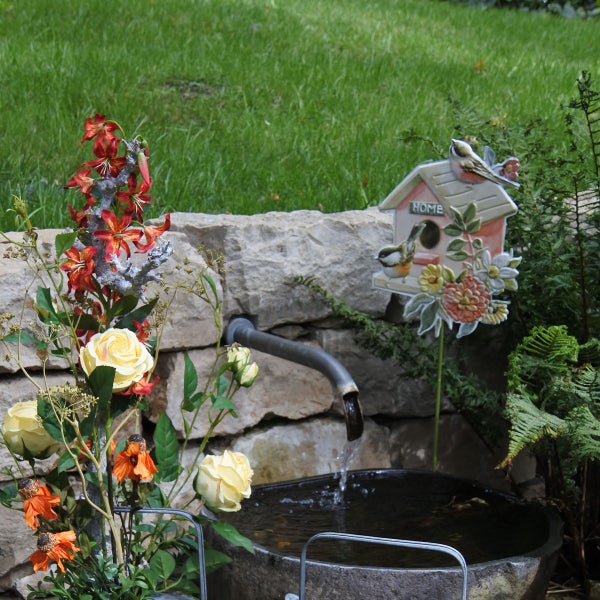 Dekorative Gartenszene mit Wasserhahn, Wasserbecken, Blumenarrangement und Vogelhaus-Dekoration.
