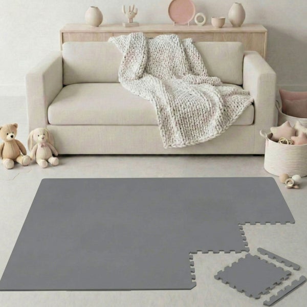 Graue Puzzlematte aus Schaumstoff mit verzahnten Kanten in einem hellen Raum vor einem Sofa mit Strickdecke und Spielzeug.