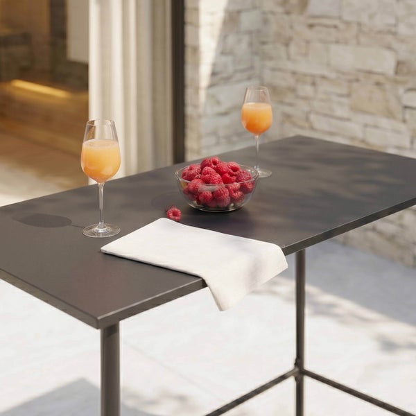 Dunkler Stehtisch aus Metall im Außenbereich mit zwei Gläsern Saft, einer Schale Himbeeren und einer weißen Serviette vor einer Steinmauer.