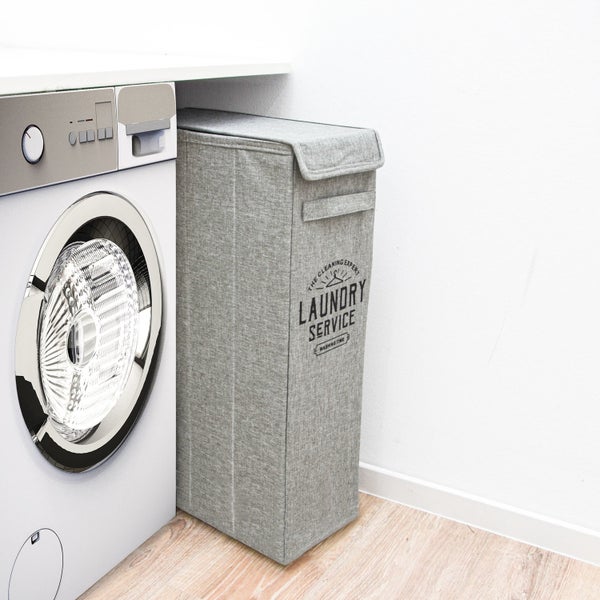 Schmaler Wäschekorb aus Stoff mit Deckel und Aufdruck Laundry Service neben einer Waschmaschine in einer hellen Waschküche.