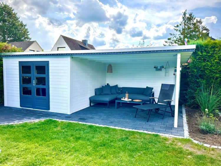 Weißes Gartenhaus aus Holz mit Pultdach, blauer Doppeltür und überdachtem Loungebereich mit Gartenmöbeln auf gepflasterter Fläche.