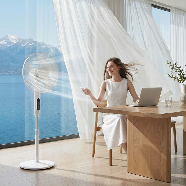 Weißer Standventilator mit Fernbedienung in hellem Raum mit Frau am Tisch, Laptop und Seeblick.