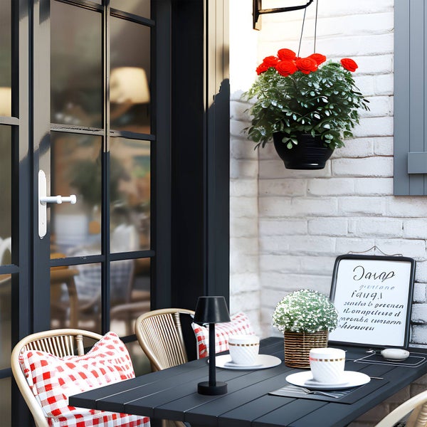 Schwarzer Gartentisch mit Stühlen, rot-weiß karierten Kissen und Lampe. Tassen und Pflanzen auf dem Tisch. Rote Rosen an weißer Ziegelwand.