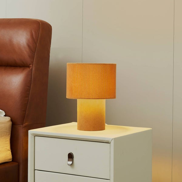 Moderne Tischleuchte mit geriffeltem Schirm und Fuß in Senfgelb auf einem hellen Nachttisch neben einem braunen Lederkopfteil.