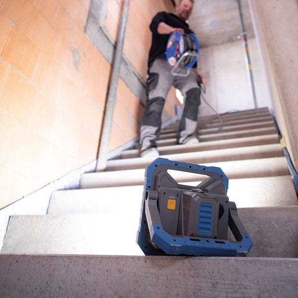 Baustellenleuchte auf einer Treppe mit einer Person im Hintergrund, die eine Kabeltrommel trägt.