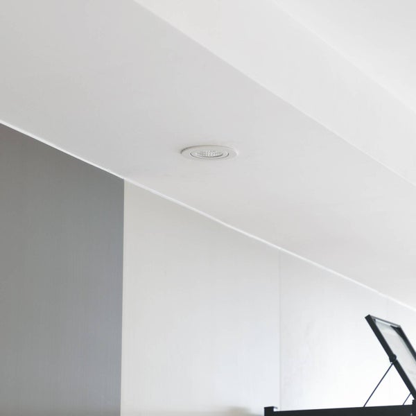 Ein runder LED-Einbaustrahler in einer weißen Decke über einer grauen und weißen Wand in einem modern gestalteten Raum.