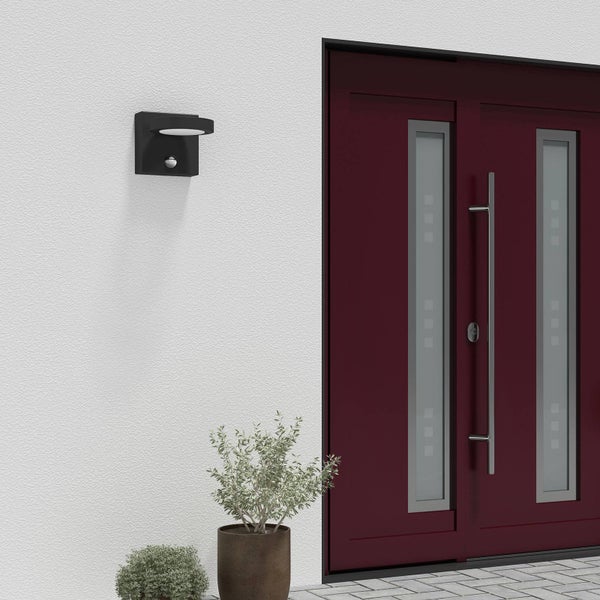 Moderne schwarze Außenwandleuchte mit Bewegungsmelder, montiert an einer weißen Wand neben einer dunkelroten Haustür.