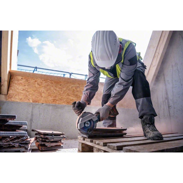 Bauarbeiter in Schutzkleidung schneidet Dachziegel mit einem Winkelschleifer auf einer Palette auf einer hellen Baustelle.