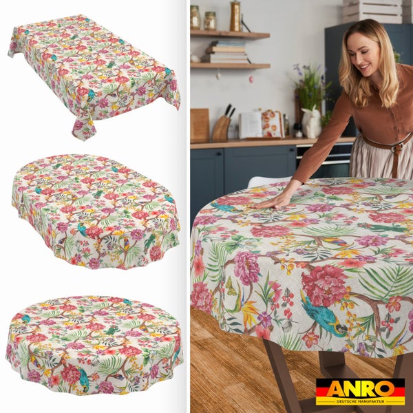 Tischdecke mit Blumen- und Papageienmuster in rechteckiger, ovaler und runder Form. Frau deckt Holztisch in heller Küche. ANRO Logo.