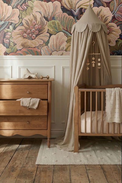 Eingerichtetes Kinderzimmer mit Holzkommode, Babybett mit Betthimmel und floraler Tapete über weißer Wandvertäfelung.