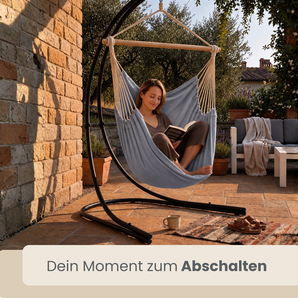 Frau liest Buch in einem Hängesessel mit Metallgestell auf einer Terrasse im Garten.