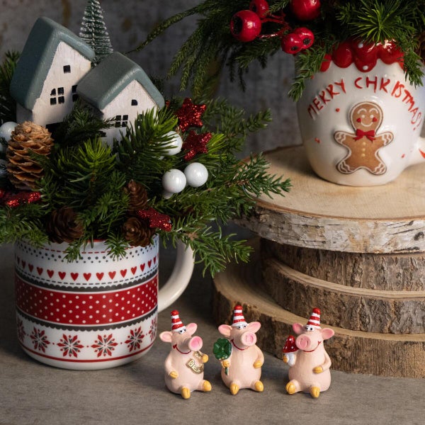 Weihnachtliche Tischdekoration mit zwei Tassen als Pflanzgefäße, geschmückt mit Tannenreisig, Miniaturhäusern, Zapfen und drei Glücksschweinchen.