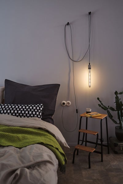 Schlafzimmer mit Bett, hängender Vintage-Glühbirne an der Wand und Tritthocker aus Holz als Beistelltisch.