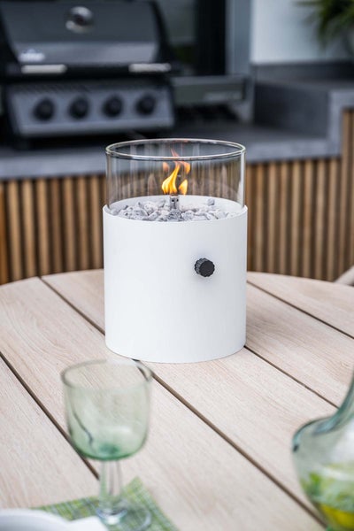 Moderne Tisch-Gasfeuerstelle mit weißem Sockel, Glaszylinder und Lavasteinen auf einem hellen Holztisch im hellen Außenbereich.