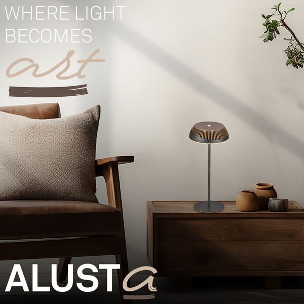 Moderne Tischleuchte auf einer Holzkommode neben einem Sessel mit Kissen im hellen Wohnzimmer. Markenschriftzug ALUSTA.