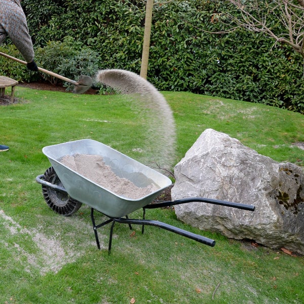 Eine Person schaufelt Sand in eine Metallschubkarre auf einer Rasenfläche im Garten, neben einem großen Naturstein und einer Hecke.