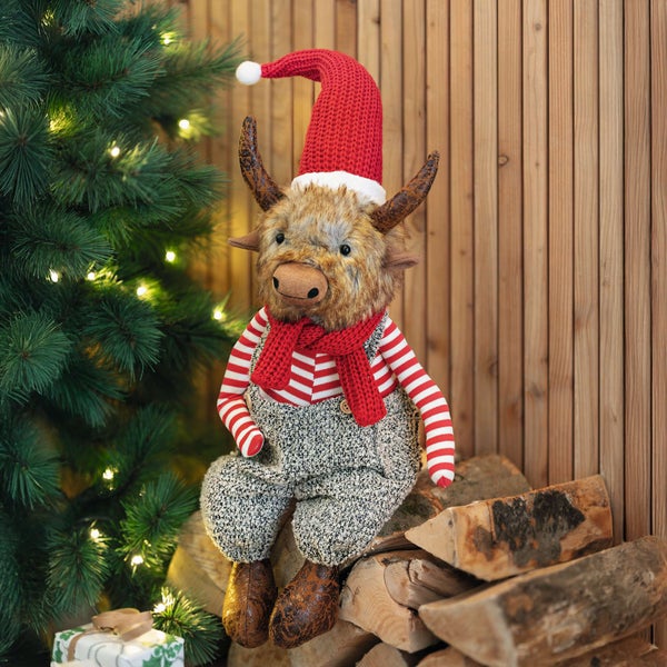Dekorative Stoffpuppe als Ochse mit Weihnachtsmütze, Schal und Overall neben einem Weihnachtsbaum