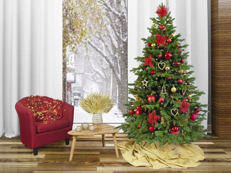 Festlich geschmückter Weihnachtsbaum mit Kugeln und Sternen neben einem Sessel und einem Beistelltisch vor einem Fenster mit Winterlandschaft.