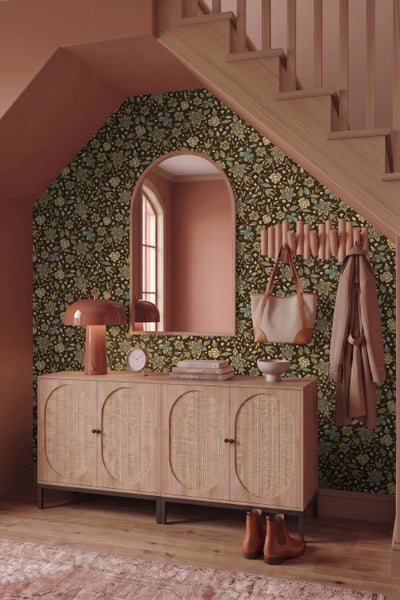 Flur mit floral gemusterter Tapete, Sideboard aus Holz mit Rattantüren, Bogen-Spiegel und Garderobe unter einer Treppe.