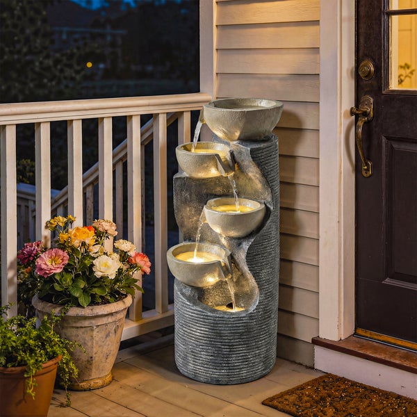 Mehrstufiger Gartenbrunnen in Steinoptik mit vier beleuchteten Schalen auf einer Terrasse neben Blumenkübeln.