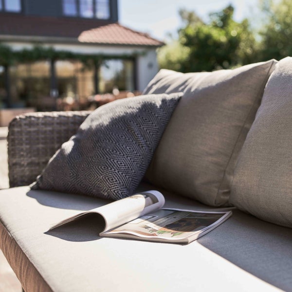 Outdoor-Lounge-Sofa aus Polyrattan mit Sitz- und Rückenkissen sowie einem Dekokissen mit Rautenmuster auf einer sonnigen Terrasse.