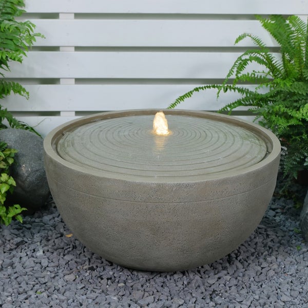 Runder Gartenbrunnen in Schalenform mit Steinoptik, Wellenstruktur und beleuchtetem Wasserspiel auf Kies zwischen Pflanzen.
