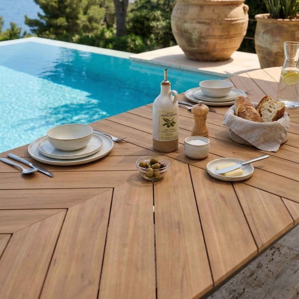 Gedeckter Gartentisch aus Holz mit Fischgrätenmuster an einem hellen Pool, dekoriert mit Geschirr, Brot und Olivenöl in mediterranem Stil.