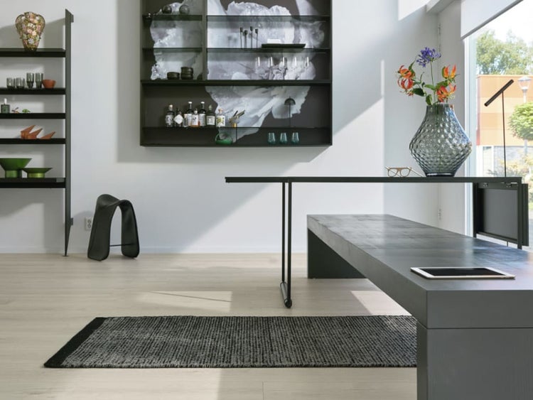 Modernes Esszimmer mit einem langen Tisch, einer Sitzbank, Regalen und einem dunklen Teppich auf hellem Boden in einem hellen Raum.