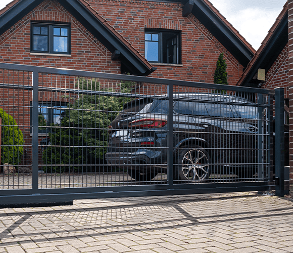 Modernes Schiebetor aus Doppelstabmatten in einer Hauseinfahrt aus Steinpflaster vor einem Backsteinhaus mit geparktem Auto in heller Atmosphäre.