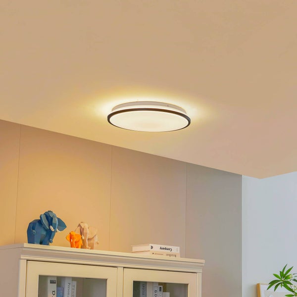 Moderne runde LED-Deckenleuchte mit schwarzem Rand in einem hellen Wohnraum über einem Schrank mit Dekorationsartikeln.