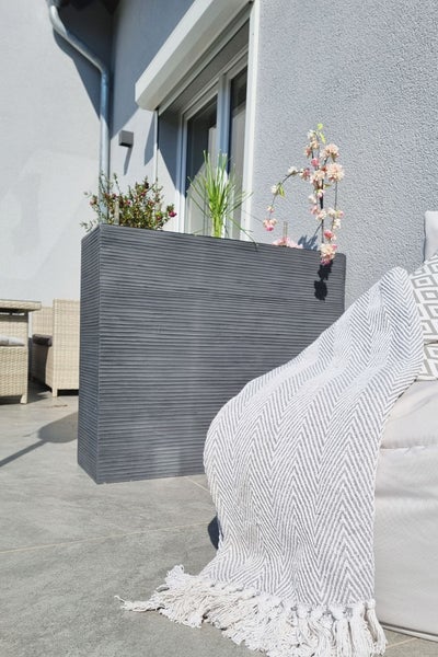 Hoher anthrazitfarbener Pflanztrog mit Rillenstruktur auf einer Terrasse, bepflanzt mit verschiedenen Gartenpflanzen neben einem Loungesofa.