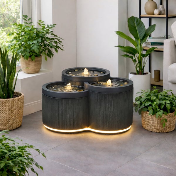 Zimmerbrunnen mit drei Säulen in dunkler Optik mit beleuchteten Fontänen und beleuchtetem Sockel, umgeben von Zimmerpflanzen.