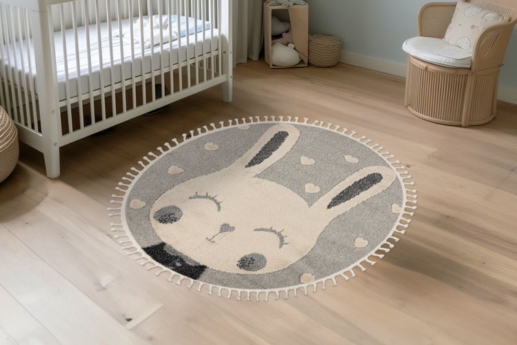 Runder Kinderteppich mit Hasenmotiv und Fransenrand in einem hellen Kinderzimmer mit Holzboden und Gitterbett.