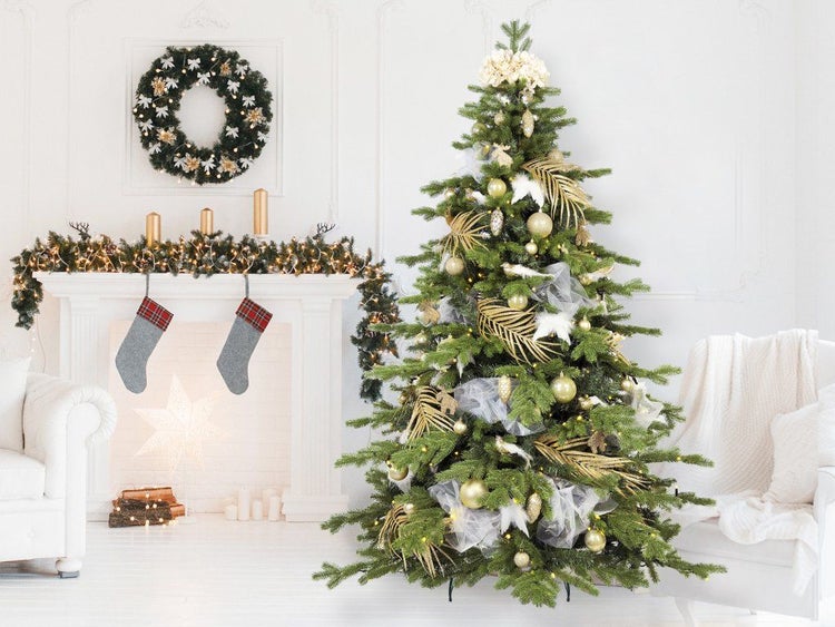Festlich dekorierter künstlicher Weihnachtsbaum mit Goldschmuck und Lichtern in einem hellen Wohnzimmer mit Kamin und Weihnachtsdekoration.