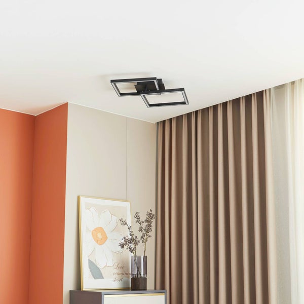 Moderne schwarze LED-Deckenleuchte mit zwei überlappenden quadratischen Rahmen in einem hellen, aufgeräumten Zimmer mit Vorhängen und Sideboard.