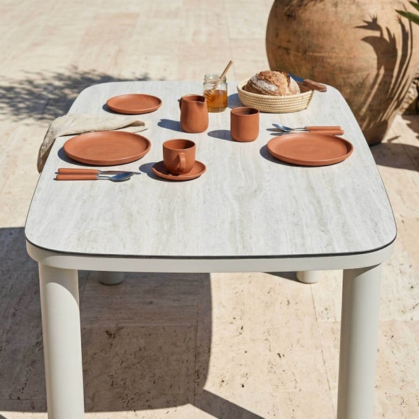Moderner weißer Garten-Esstisch mit Tischplatte in Steinoptik, gedeckt mit terracottafarbenem Geschirr auf einer sonnigen Steinterrasse.