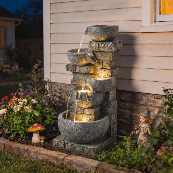 Mehrstöckiger Gartenbrunnen in Steinoptik mit warmem LED-Licht auf einer Terrasse am Abend, im Hintergrund ein Paar auf Loungemöbeln.