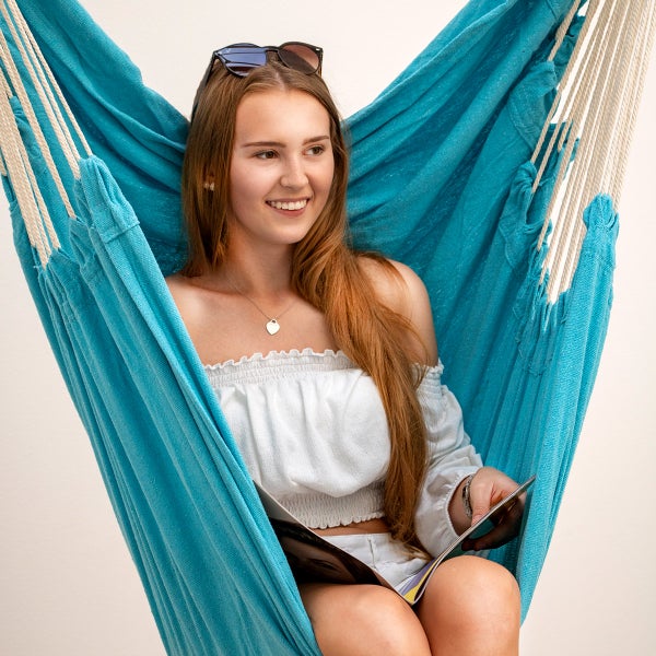 Eine junge Frau entspannt sich in einem blauen Hängesessel aus Stoff und liest eine Zeitschrift.