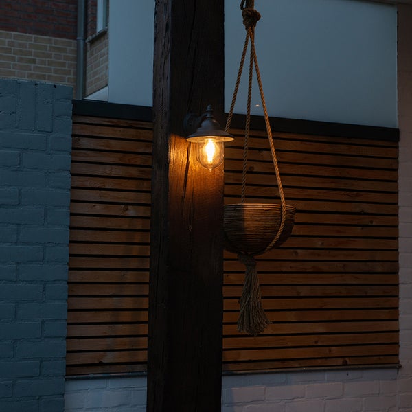 Außenwandleuchte im Laternenstil an einem dunklen Holzbalken, beleuchtet, neben einer hängenden Pflanzschale vor einem Holzzaun.
