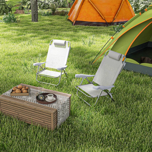 Zwei klappbare Campingstühle mit Kopfstützen neben einem Holztisch und Zelten auf einer Rasenfläche.