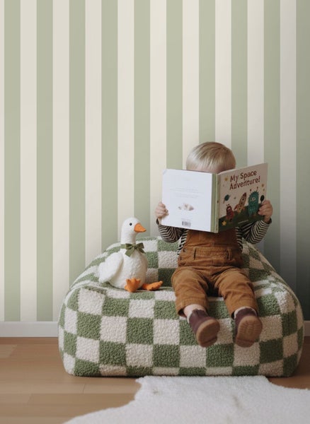 Kind auf einem karierten Sitzsack liest ein Buch vor einer Wand mit vertikal gestreifter Tapete in einem hellen Zimmer.